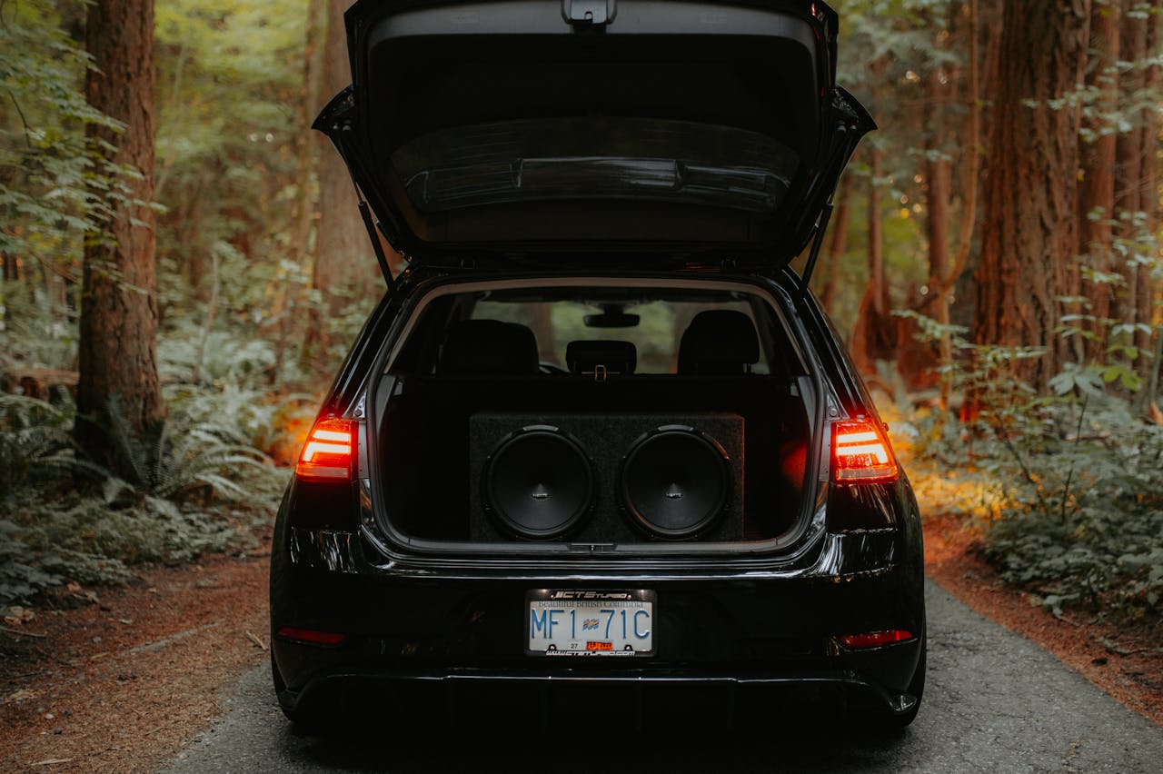 A car parked in a forest with its trunk open, revealing large speakers inside.